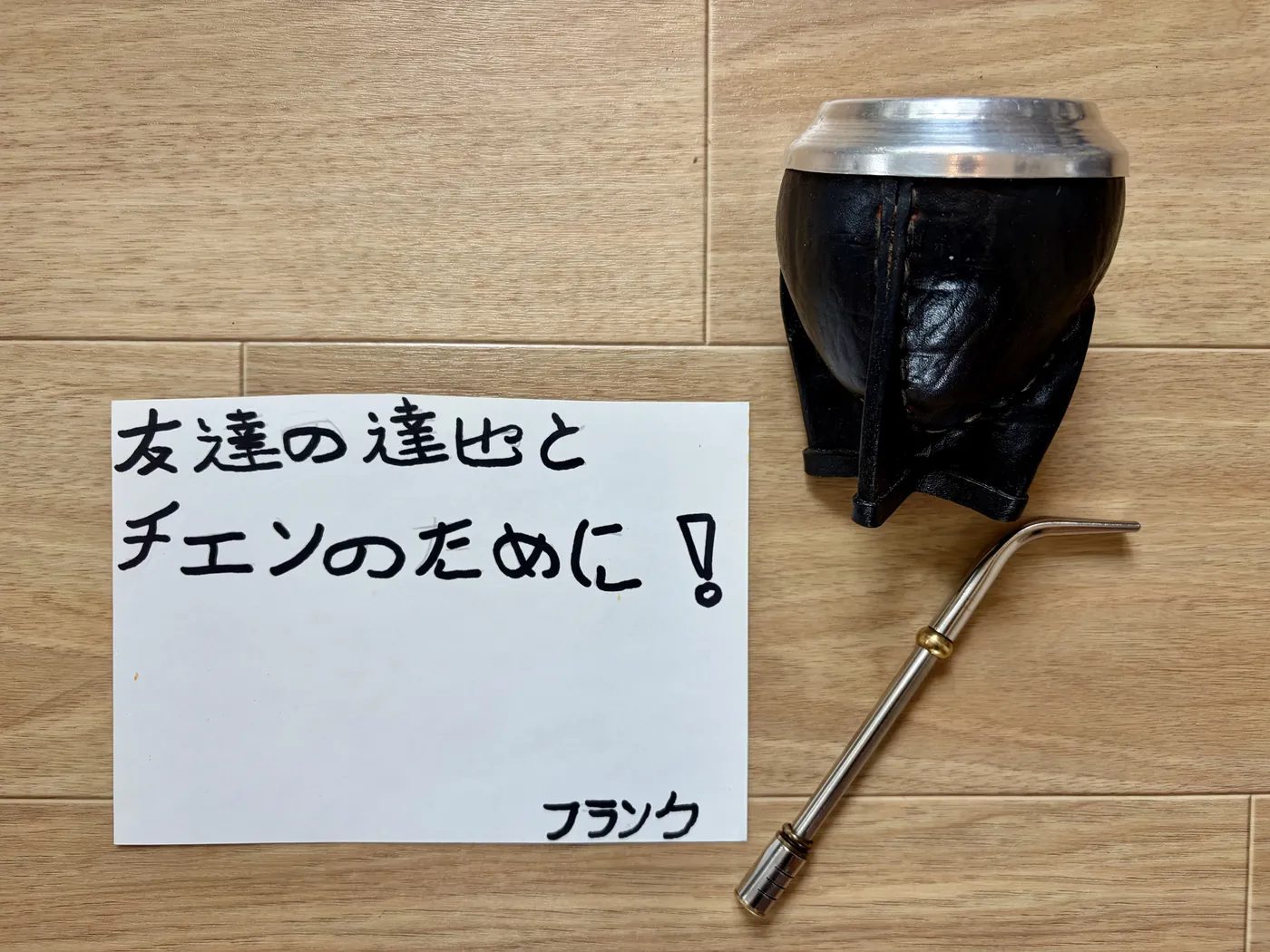 木の床に置かれた、革を巻いたひょうたん製のマテ壺と金属のボンビーシャ。手書きのメッセージカードが添えられている