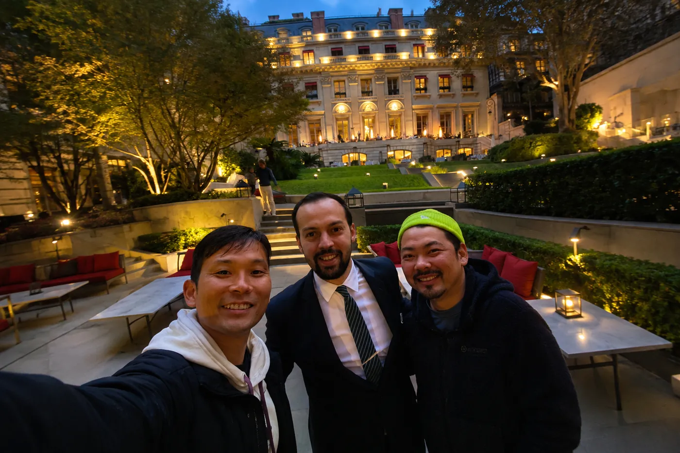 夜の庭園。ライトアップされた建物を背に、スーツ姿の男性を挟んで三人が並んでいる